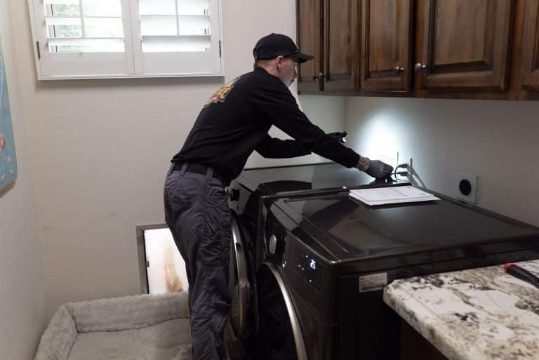 Technician using a flashlight to check behind a washer and dryer for mouse entry points